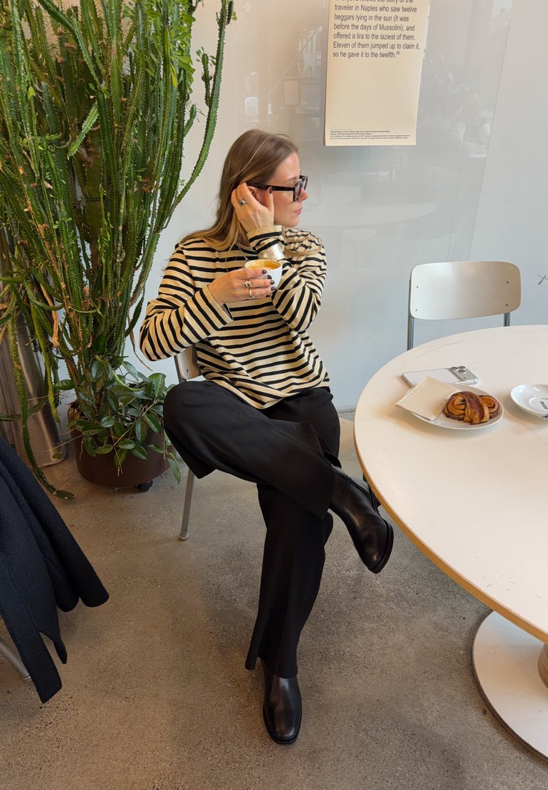 Kvinde iført sort og hvidt stribet sweater og briller sidder med benene over kors og holder en kaffekop ved siden af et hvidt bord med kager.