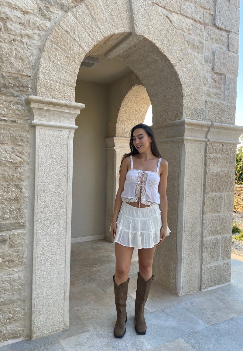Λευκή crop top με δαντέλα, με κόμπο μπροστά, στρωμένη λευκή φούστα με ρίγες και καφέ καουμπόικες μπότες μέχρι το γόνατο, μπροστά από έναν πέτρινο αψίδα.
