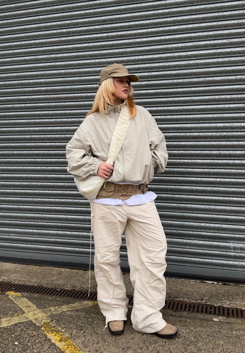 Personne portant une veste beige, un pantalon large, une casquette et des bottes, debout devant un volet métallique, les mains dans les poches et un sac bandoulière.