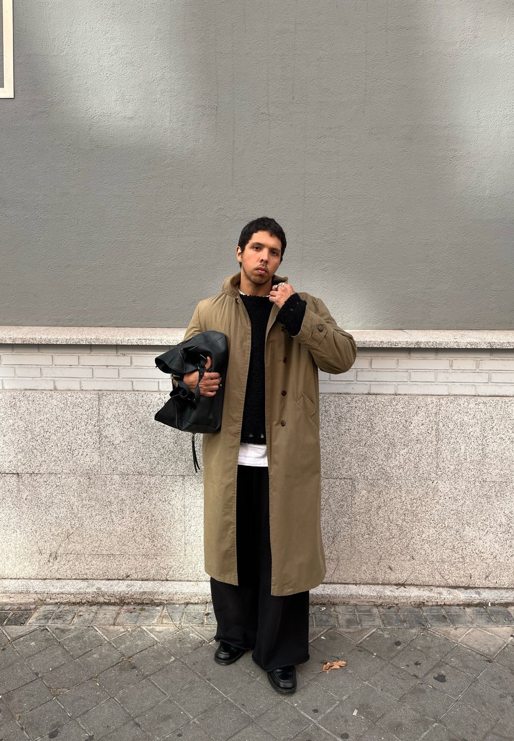 Μακρύ καφέ παλτό πάνω από μαύρο πουλόβερ, μαύρο παντελόνι με φαρδιά γραμμή και μαύρες παπούτσια; κρατά μια μαύρη δερμάτινη τσάντα; στέκεται μπροστά σε γκρίζο τοίχο.