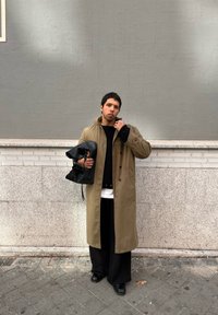 Μακρύ καφέ παλτό πάνω από μαύρο πουλόβερ, μαύρο παντελόνι με φαρδιά γραμμή και μαύρες παπούτσια; κρατά μια μαύρη δερμάτινη τσάντα; στέκεται μπροστά σε γκρίζο τοίχο.