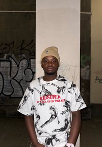 Weiss T-Shirt mit schwarzen künstlerischen Figuren, rotem "KIDSUPER"-Text, kombiniert mit einer beigen Mütze. Der Hintergrund zeigt Graffiti an einer grauen Wand.