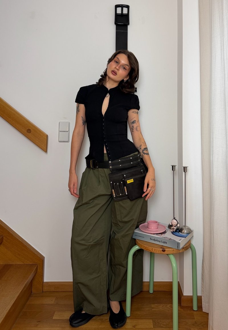 Joven mujer con tatuajes, vestida con top negro y pantalones verdes anchos, de pie en el interior cerca de una escalera y una pequeña mesa con libros y tazas.