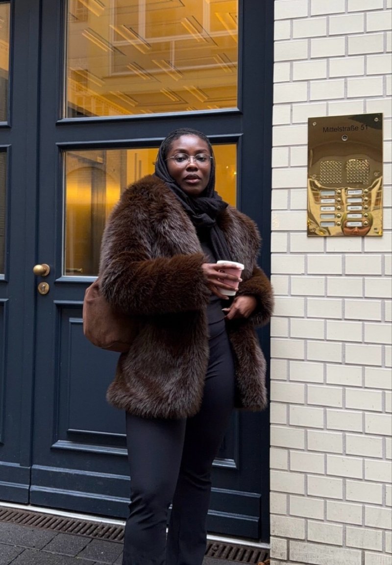 Manteau en fausse fourrure marron, écharpe noire, pantalon noir, tenant une tasse blanche, debout devant un mur de briques texturé et un interphone de porte.
