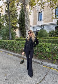 Giacca di tweed nera con bottoni dorati, top nero e pantaloni neri svasati. Accessorio: piccola borsa a pois neri. Ambientazione all'aperto con vegetazione.