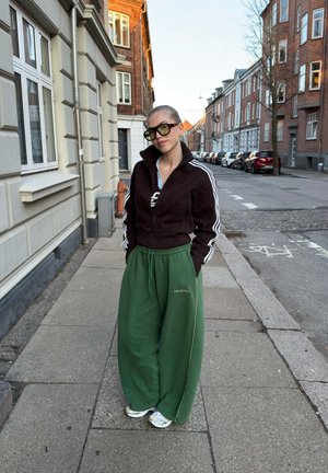 Persona que lleva puestos pantalones verdes de gran tamaño, chaqueta negra con cremallera y rayas blancas, y gafas de sol grandes, de pie en una acera urbana.