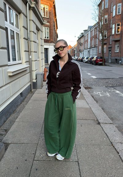 Persona que lleva puestos pantalones verdes de gran tamaño, chaqueta negra con cremallera y rayas blancas, y gafas de sol grandes, de pie en una acera urbana.