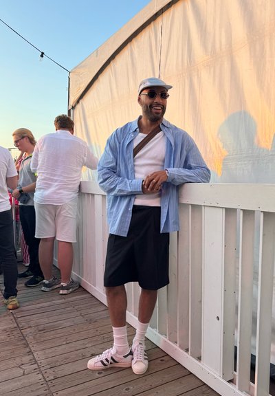 Camisa de rayas azul claro de manga corta, camiseta de tirantes blanca, pantalones cortos negros, calcetines tobilleros blancos y zapatillas blancas con acentos negros. Entorno interior.