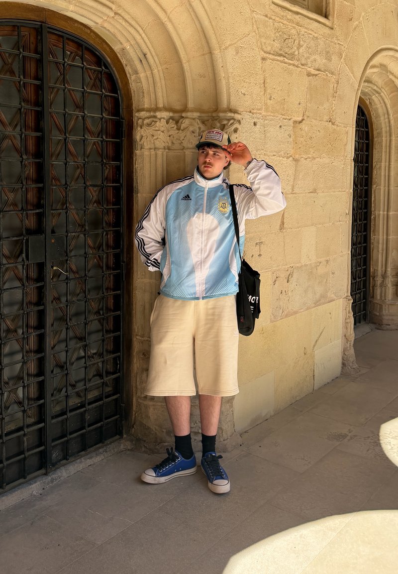 Young man in light blue and white sports jacket and beige shorts adjusting cap, standing by ornate stone wall and black barred door.
