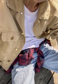 Casaco de veludo cotelê bege com botões dourados, sobre uma t-shirt branca. Camisa xadrez vermelha e preta amarrada à cintura, combinada com jeans azul-claro.