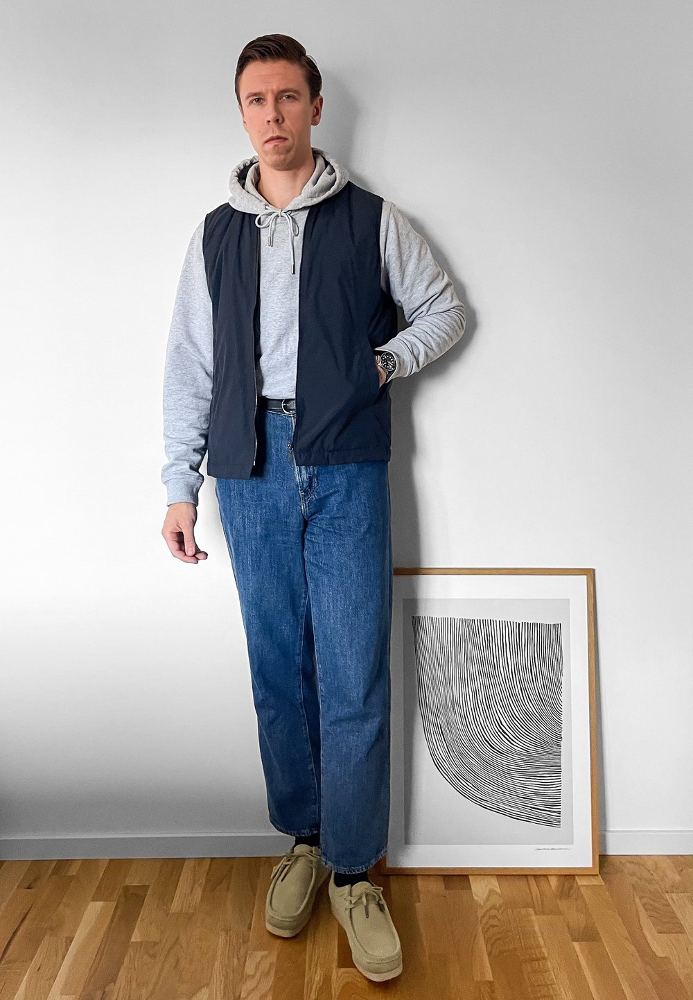 Ležérní outfit se skládá z námořnického bezrukávového vesty přes šedou mikinu, modrých denimových džínů a béžových semišových bot. Vedle je rámovaný obraz.