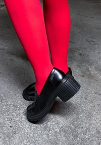 Πόδια με φωτεινές κόκκινες καλσόν που φορούν μαύρα χοντρά μοκασίνια με πάτο πλατφόρμα σε γκρι τσιμεντένιο δάπεδο.