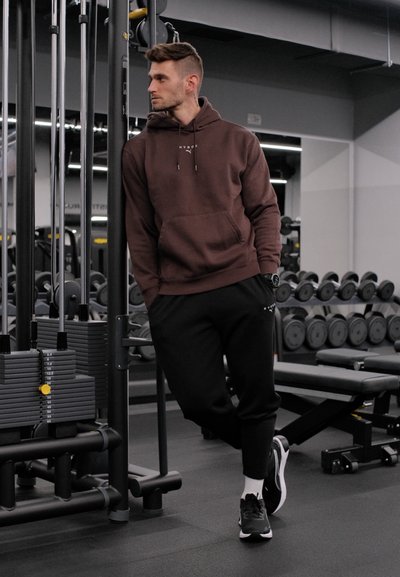 Hombre con sudadera marrón con capucha y pantalones negros apoyado en una máquina de pesas de gimnasio, mancuernas y banco visibles en un entorno moderno de gimnasio.