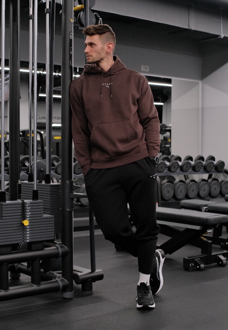 Homme en sweat à capuche marron et pantalon noir appuyé contre une machine de musculation, haltères et banc visibles dans une salle de sport moderne.