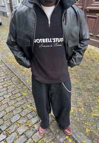 Black leather jacket, black knitted sweater with white text, loose black trousers, and brown shoes. Outdoor setting with cobbled ground.