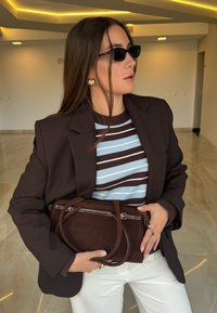 Blazer marrone sopra una maglia a righe blu e marrone; tiene una piccola borsa marrone con doppi manici. Capelli lunghi, occhiali da sole neri, pantaloni neutri.