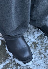 Bottes imperméables noires à finition lisse, debout sur un sol enneigé, associées à un pantalon en tissu gris, montrant une légère humidité sur la surface des bottes.