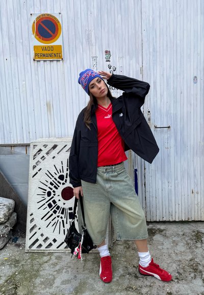 Joven con camiseta roja de Puma, chaqueta negra, pantalones cortos vaqueros anchos, zapatillas rojas de Puma y un gorro azul posando junto a una puerta metálica blanca con un cartel de no estacionar.