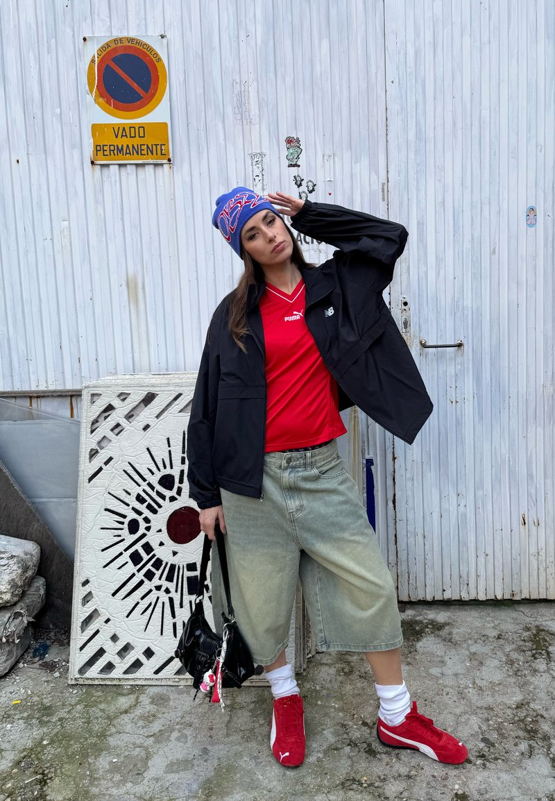 Ung kvinne iført rød Puma-skjorte, svart jakke, vide denimshorts, røde Puma-sneakers og en blå lue som poserer ved en hvit metalldør med et skilt for parkering forbudt.