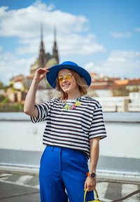 Camiseta listrada em preto e branco, calças azuis vibrantes, chapéu azul, óculos de sol amarelos e um colar floral colorido. Detalhe da bolsa amarela visível.