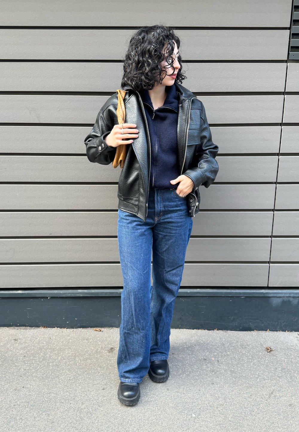 Veste en cuir noir à finition texturée, sweat à capuche bleu marine, jean évasé bleu et chaussures à plateforme noires. Objet marron tenu sous le bras gauche.