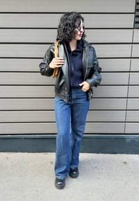 Veste en cuir noir à finition texturée, sweat à capuche bleu marine, jean évasé bleu et chaussures à plateforme noires. Objet marron tenu sous le bras gauche.