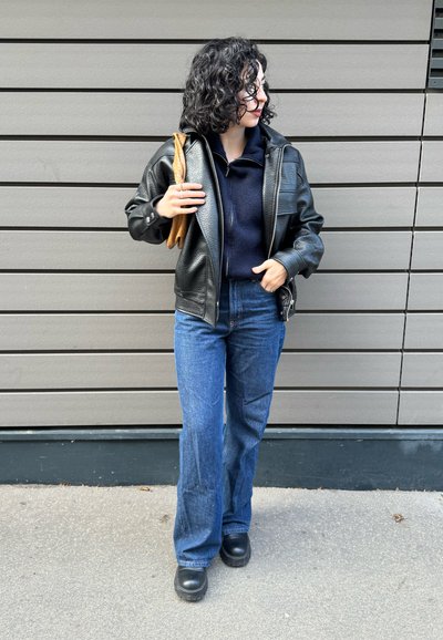 Chaqueta de cuero negra con acabado texturizado, sudadera con capucha azul marino, vaqueros acampanados azules y zapatos plataforma negros. Objeto marrón sostenido bajo el brazo izquierdo.