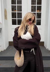 Ξανθιά γυναίκα με γυαλιά ηλίου, σκούρο παλτό, λευκό πουλόβερ και κρατώντας μία μπεζ  τσάντα, στέκεται με σταυρωμένα τα χέρια μπροστά σε λευκά σκαλοπάτια πόρτας.