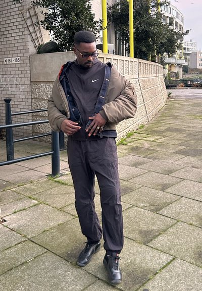 Chaqueta de plumón verde oliva sobre un sudadera negra de Nike, combinada con un pantalón negro holgado y zapatos negros. Entorno urbano con una pared de ladrillo.