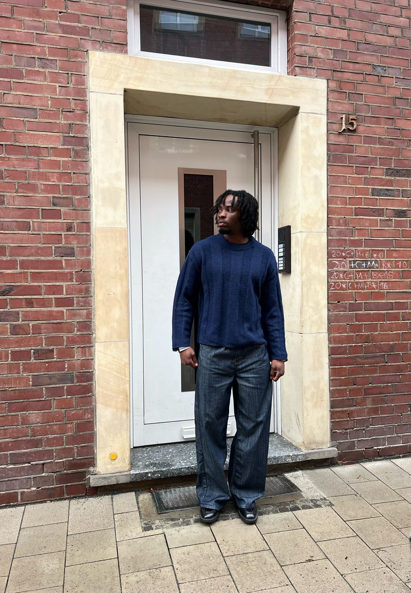 Homme aux dreadlocks portant un pull bleu marine et un jean large, debout devant une porte blanche encadrée de pierre beige sur un mur en briques rouges.
