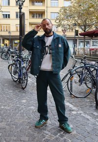 Zöld dzseki szürke pulóver felett, melyen "DEEP" felirat található, zöld nadrággal és zöld sportcipőkkel párosítva. Köves utca és kerékpárok a háttérben.
