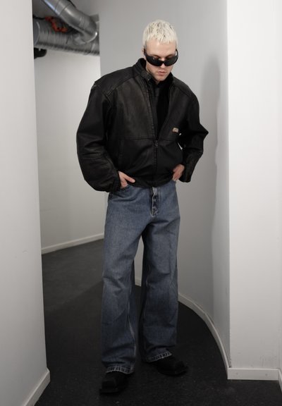 Hombre con cabello decolorado que lleva una chaqueta de cuero negra, gafas de sol oscuras, jeans azules holgados y zapatos negros, de pie en un pasillo estrecho.