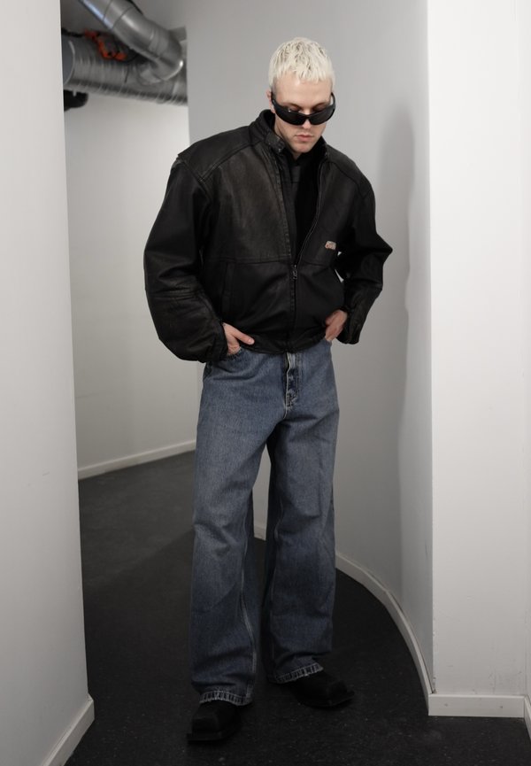 Man met gebleekt haar, gekleed in een zwart leren jack, donkere zonnebril, losse blauwe jeans en zwarte schoenen, staande in een smalle gang.