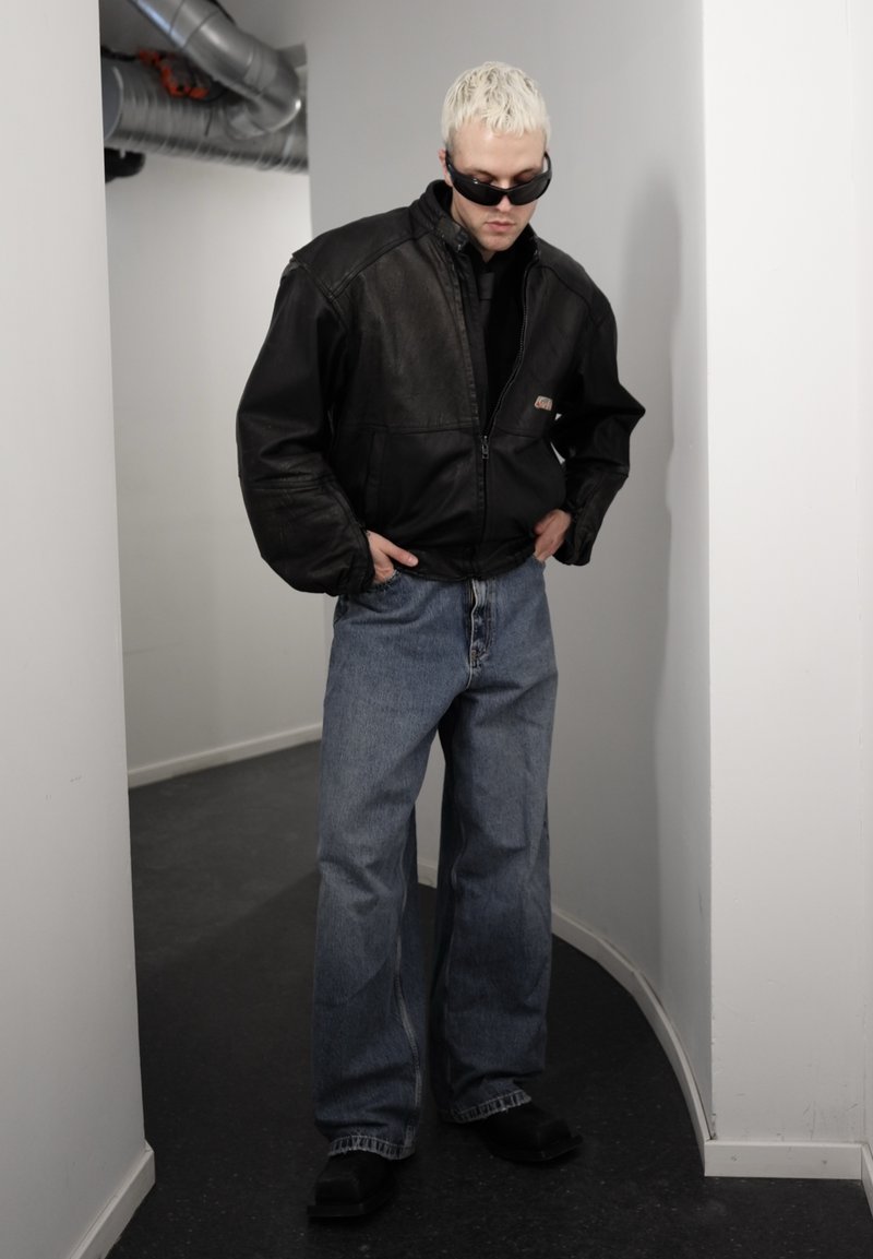 Homme aux cheveux décolorés portant une veste en cuir noire, des lunettes de soleil foncées, un jean bleu large et des chaussures noires, debout dans un couloir étroit.