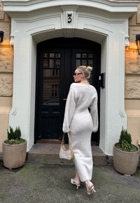 Γυναίκα με μακρύ λευκό πλεκτό φόρεμα και τακούνια κρατώντας μια μπεζ τσάντα, στέκεται μπροστά από μια μαύρη πόρτα με τον αρ ιθμό 3 πάνω της.