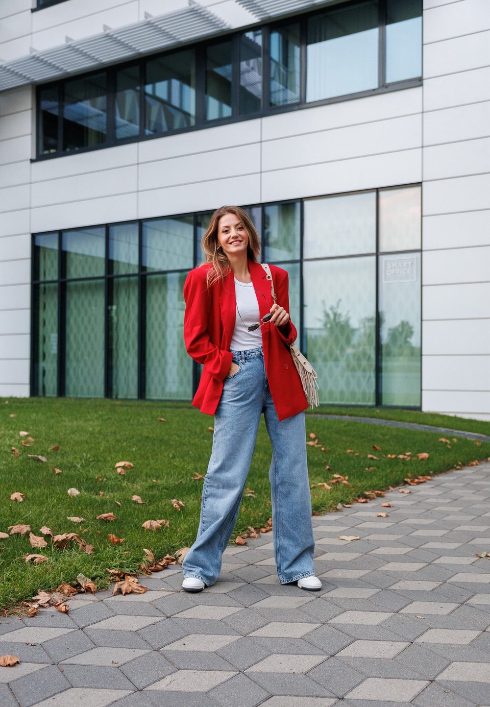 Κόκκινο υπερμεγέθες σακάκι, λευκό T-shirt και φαρδιά μπλε τζιν; λευκά αθλητικά και τσάντα με φούντες; στέκεται σε μια πλακόστρωτη περιοχή με χόρτο και πεσμένα φύλλα.