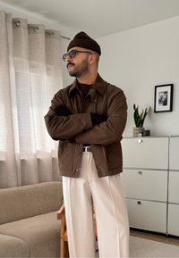 Veste marron zippée avec un col en velours côtelé, gants noirs, pantalon large crème et bonnet marron ; debout dans une pièce bien éclairée.