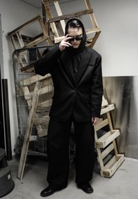 Traje negro de gran tamaño con una sudadera con capucha, gafas de sol negras y uñas largas. El fondo presenta cajas de madera y una pared metálica.