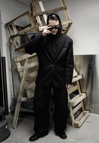 Traje negro de gran tamaño con una sudadera con capucha, gafas de sol negras y uñas largas. El fondo presenta cajas de madera y una pared metálica.