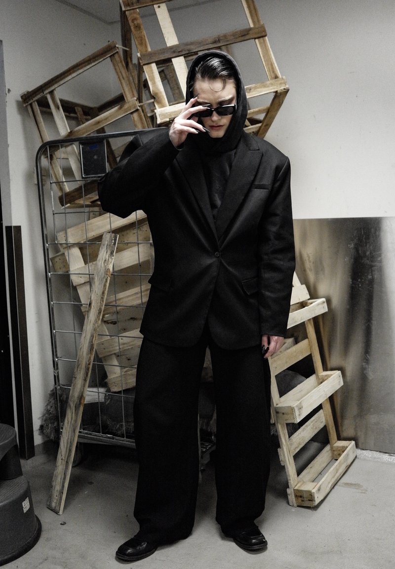 Schwarzer übergroßer Anzug mit Kapuzenoberteil, schwarzen Sonnenbrillen und langen Nägeln. Im Hintergrund sind Holzkisten und eine metallische Wand zu sehen.