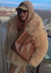 Personne portant un manteau en fourrure beige avec capuche, lunettes de soleil et casquette, tenant un grand sac en cuir marron sur un toit avec une vue sur la ville en arrière-plan.