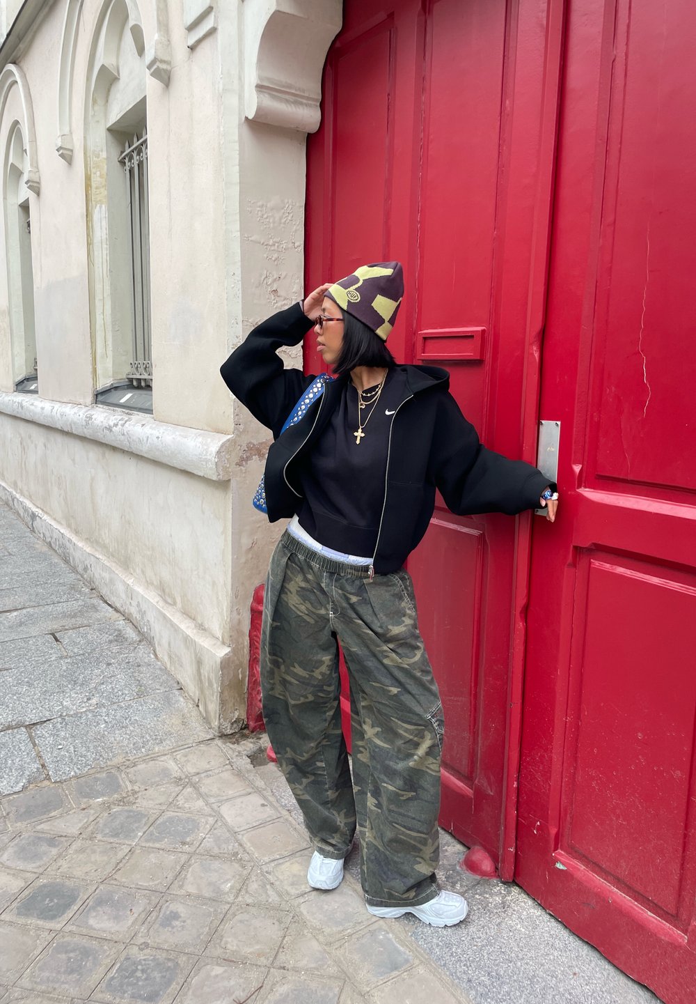 Sweat à capuche noir avec fermeture éclair, pantalon camouflage, bonnet coloré et baskets blanches. Appuyé contre une porte rouge dans une rue pavée.