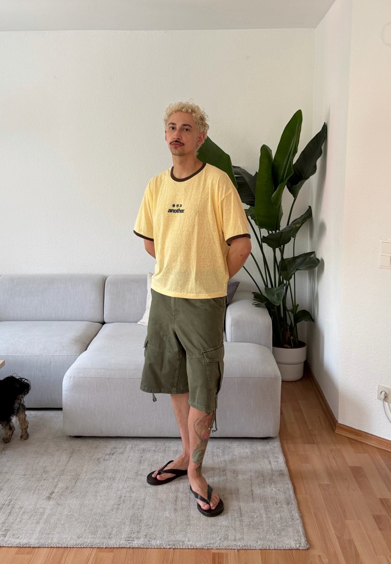 Homme aux cheveux blonds bouclés, T-shirt jaune, short vert et tongs, debout dans le salon près d'un canapé gris et d'une grande plante en pot.