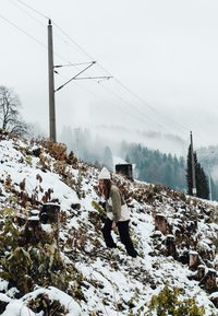Person som klatrer opp en snødekt skråning med vegetasjon og trestubber. En strømstolpe står i bakgrunnen midt i et tåkete, fjellandskap.