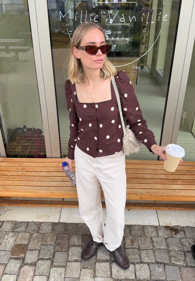 Camisa de botones marrón con estampado de lunares, pantalones anchos blancos, zapatos marrones, sosteniendo una taza de café y una botella de agua, de pie cerca de una puerta de cristal.