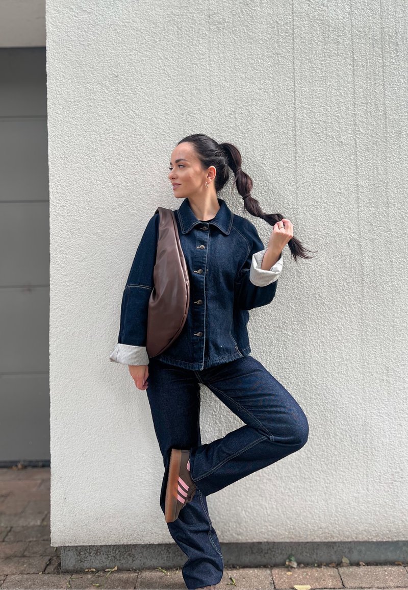Casaco de ganga escura com botões, punhos forrados a creme; jeans combinando e mala de crossbody castanha; sapatos com riscas rosas; fundo de parede texturizada.