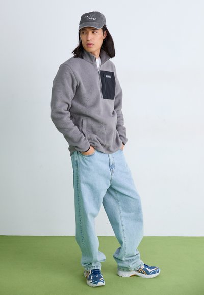 Sudadera de forro polar gris con un bolsillo en el pecho negro, combinada con jeans holgados de color azul claro y zapatillas deportivas azules. Gorras con detalle de texto.