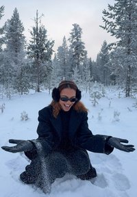 Personne portant un manteau noir, des gants, des cache-oreilles et des lunettes de soleil, agenouillée dans la neige, jetant joyeusement de la neige avec des arbres couverts de neige en arrière-plan.