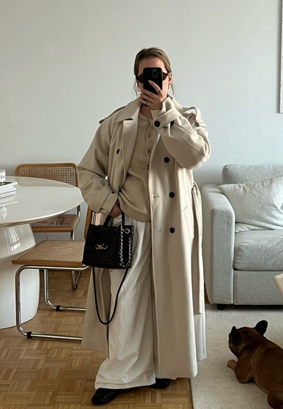 Abrigo trench beige largo con botones negros, sobre un suéter beige y pantalones de pierna ancha blancos. Bolso de mano tejido negro, gafas de sol elegantes y un perro cerca.