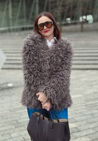 Manteau en fausse fourrure gris à texture duveteuse, porté sur une chemise blanche et un jean bleu. Un sac à main foncé complète la tenue.